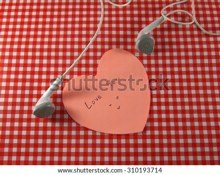 Love you and happy smiling face, handwriting on sticky note, heart shape with left, right of white earphones, tartan pattern background