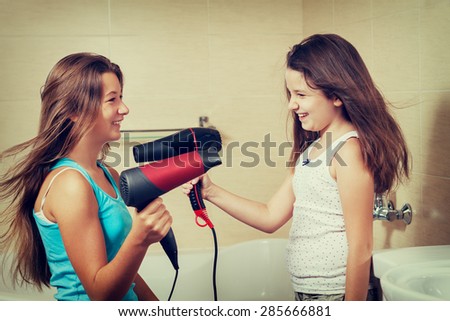 Two beautiful girls are playing in the bathroom with the hair dryer after bathing