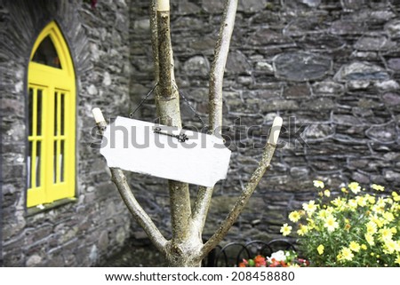 Blank white wooden board with vintage key on the tree and yellow window in stone house with flowers in the back.