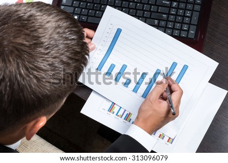 The man in a suit bent over the table with a laptop and printed charts and going to write a top view close-up
