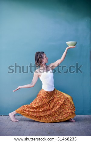 Receive. Unique woman in a dancing yoga posture offering up a beautiful ceramic bowl against the backdrop of a blue wall.