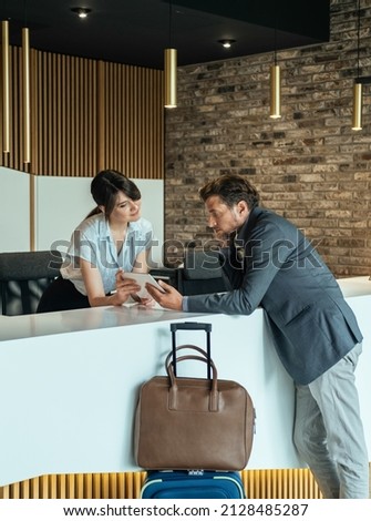 Businessman Using Digital Tablet to Checking In at Hotel. 
Businessman with luggage standing at hotel reception and filling in registration forms on digital tablet with assistance of female concierge. Foto stock © 