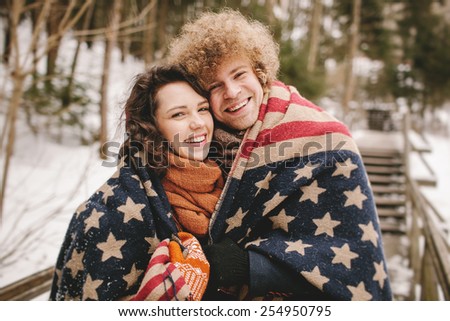 Young happy hipster couple covering under stars and stripes rug outdoors in winter park