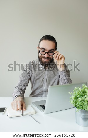 Smiling man with beard in glasses taking notes with laptop and notepad looking in camera