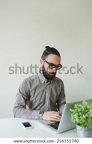 Busy man with beard in glasses typing on laptop with smartphone on the table