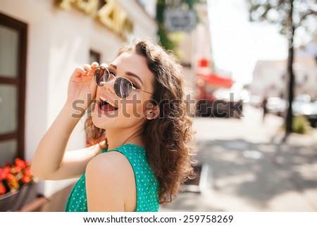 Young woman in round sunglasses and dress with curly hair smiling over the shoulder in the city street