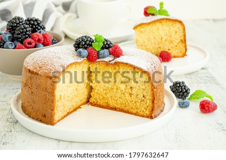 Homemade classic vanilla sponge cake or biscuit sprinkled with powdered sugar and fresh berries on top on a white plate on a light wooden background