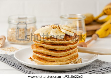 A stack of oatmeal banana pancakes with slices of fresh bananas, walnuts and honey on top with cup of tea on a white wooden background. A healthy breakfast. Copy space