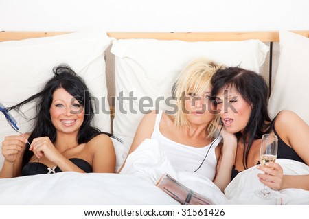 Three girls in one bed having fun in reading newspaper magazine, listening music, singing and combing hairs while celebrating bachelorette party at home living in a flat share community