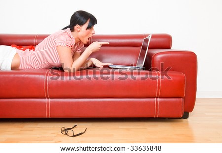 Happy surprised internet brwosing woman showing finger thumb to laptop screen while lying at home on red couch