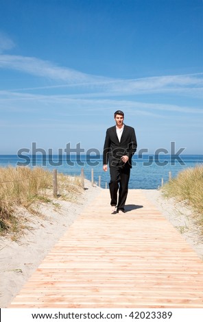 Carefree successful businessman walking with success on boardwalk on coastline with blue sky and sea wearing tie and formal clothes
