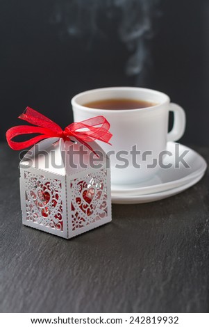 Present, white lacy box with red chocolate candies, cup od tea steaming on dark stone background