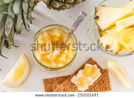 Pineapple and lemon jam in glass jar with ingredients and crisp bread on white wood background, top view