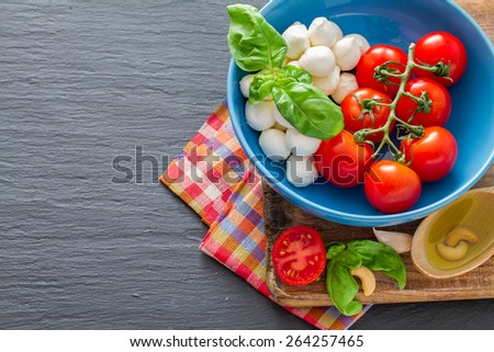 Caprese salad ingredients - cherry tomatoes, mozzarella, basil leafs in blue bowl, olive oil, nuts, chili, garlic, plaid napkin, dark stone background, top view
