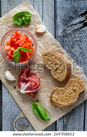 Tomato bruschetta with basil leafs, garlic, meat, cottage cheese on baking paper, rustic wood background, top view