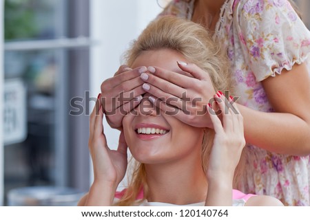 a girl with french manicure is covering eyes of her friend with hands. the friend is a beautiful young who is smiling because of the unexpected surprise