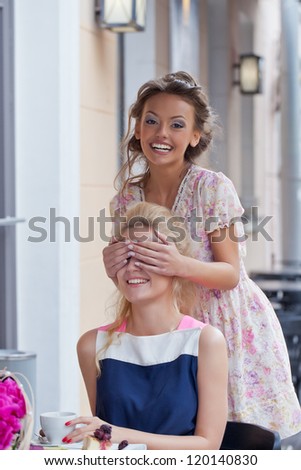 a girl with a toothy smile in summer dress is covering eyes of her beautiful friend who is drinking tea with pie