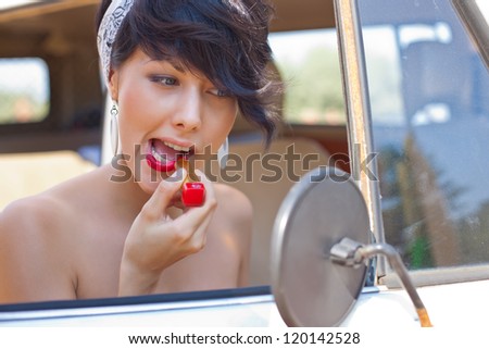 a beautiful young girl with short hair cut and blue eyes is looking into the side view mirror and is putting on red lipstick