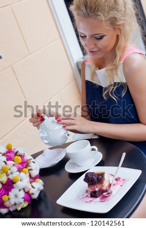 a beautiful young blond girl in summer dress at the table in pavement cafe is pouring tea from the teapot into the cup