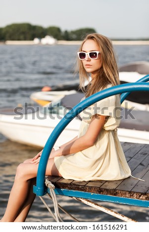 beautiful tanned girl in light summer dress and sunglasses sits under water against boats