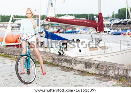 beautiful tall blonde girl in short white shorts, light blue top with cruiser bike poses for camera at sea pier against yachts
