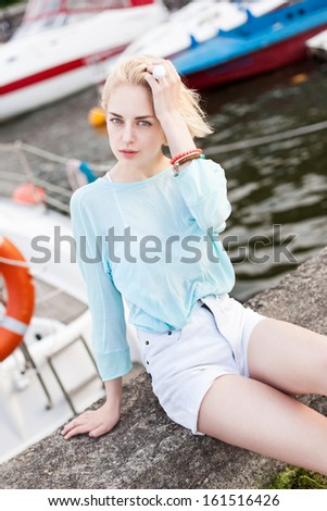 beautiful blonde girl with long legs in short white shorts, transparent light blue top sits at pier against yachts