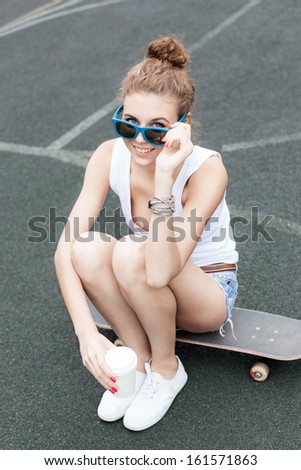 beautiful smiling girl with a white to-go cup looking over sunglasses sits on skateboard at basketball court