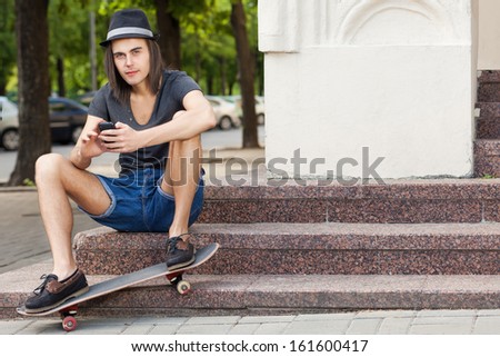 long haired handsome boy in a hat with a skateboard sits on the stairs types message on the phone