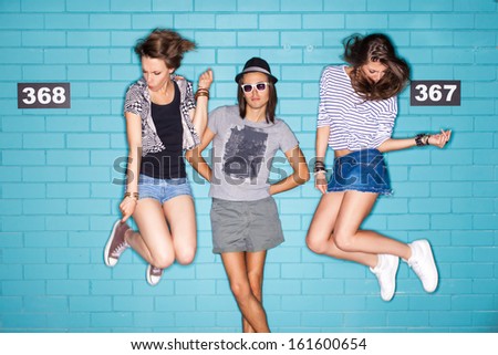 girls jump playing air invisible guitar while boy in hat and sunglasses poses for the camera in front of blue brick wall