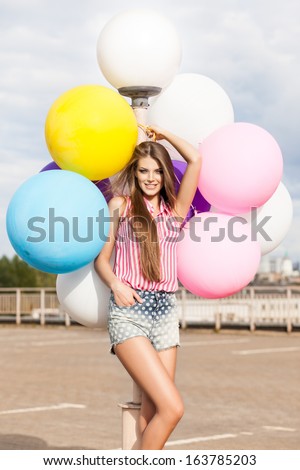 beautiful smiling lady with long silky hair in short denim shorts and sleeveless striped top leans on light-pole holding bunch of bright balloons