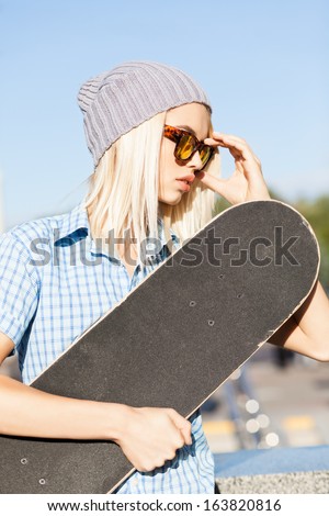 Close-up of beautiful blond girl in beanie hat skateboard adjusting her leopard sunglasses on sunny day in the street