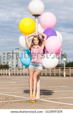 girl in short jeans shorts, sleeveless striped top and high heels leans on light-pole holding bunch of multicolored balloons in bright summer day
