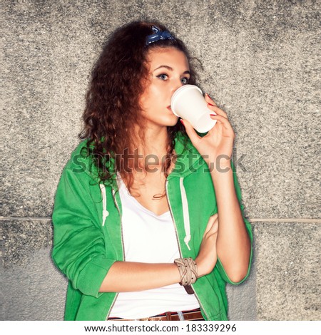 pretty young girl in green hoodie drinks from white to-go cup sits at stone wall