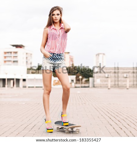 beautiful girl in short jeans shorts, sleeveless striped top and high heels stands on skateboard setting her long silky hair