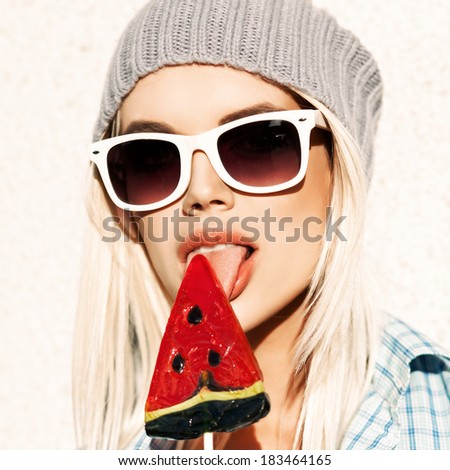 Portrait of sexy blond girl in white sunglasses and beanie hat licking watermelon lollipop with tongue