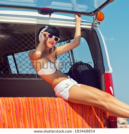 a beautiful young girl in retro look with a white swimsuit, a bandana, sunglasses and flip flops is sitting in the car trunk