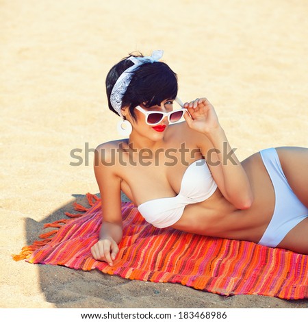 a beautiful young girl in retro look with red lips in a white swimsuit, a bandana and sunglasses is lying on the beach in the shade and winking