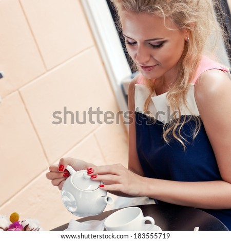 a beautiful young blond girl in summer dress at the table in pavement cafe is pouring tea from the teapot into the cup