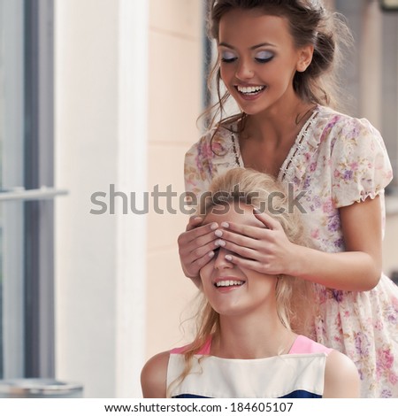 a girl with a toothy smile in summer dress is covering eyes of her beautiful friend who is eating pie with berries