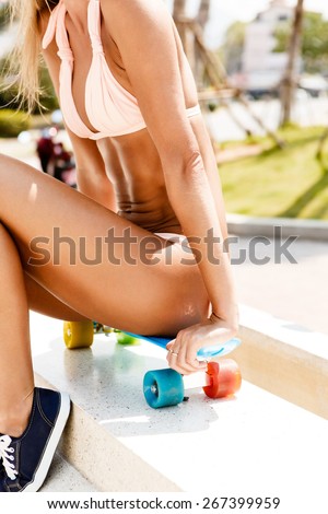 Sexy sporty tanned woman in bikini sitting on her blue penny board longboard with multi colored wheels