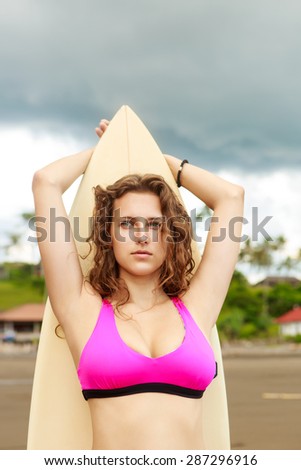 beautiful young lady with curly hair in pink bikini with surfboard standing on the background of a tropical landscape on the sea shore