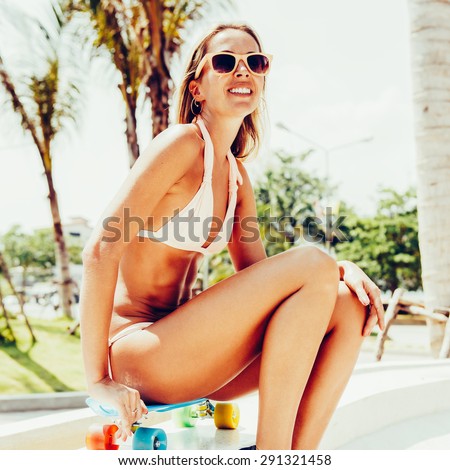 Sensual tanned woman in bikini and sunglasses  sitting on her blue penny board longboard with multi colored wheels and smiles