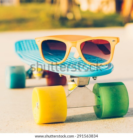 close up of the sunglasses on the blue penny board with multicolored wheels in front of the palm trees