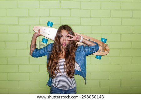beautiful long-haired lady with wooden skateboard show victory sign to the camera while poses near the green brick wall