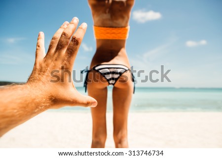 Mans hand trying to touch tanned fit butt of a sporty young beautiful female in a sexy stripped bikini on a sea shore background. Outdoor lifestyle picture on a hot sunny summer day.