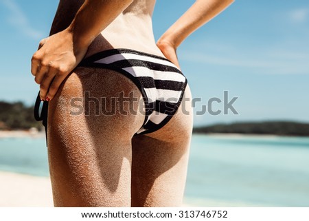 Sandy buttocks of a young beautiful sporty lady in striped black and white bikini panties on the tropical ocean shore background. Outdoor lifestyle picture on a hot sunny summer day.