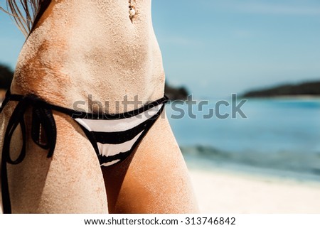 Sandy abs of a young beautiful sporty lady in striped black and white bikini panties on the tropical ocean shore background. Outdoor lifestyle picture on a hot sunny summer day.