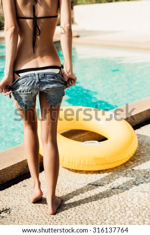 Sporty young woman with sexy athletic butt in a striped bikini with yellow inflatable swimming ring takes off her jeans shorts at the refreshing pool. Outdoor lifestyle picture on a sunny summer day.