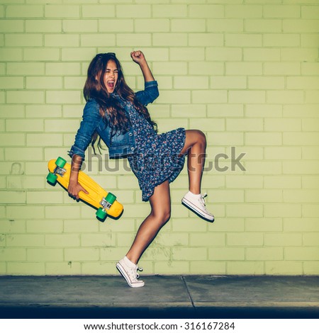 young happy beautiful long-haired brunette girl in blue dress having fun with yellow plastic penny board skateboard in front of the green brick wall