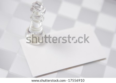 Glass king figure staying on the white blank business card (visit card), on the chessboard. Closeup.